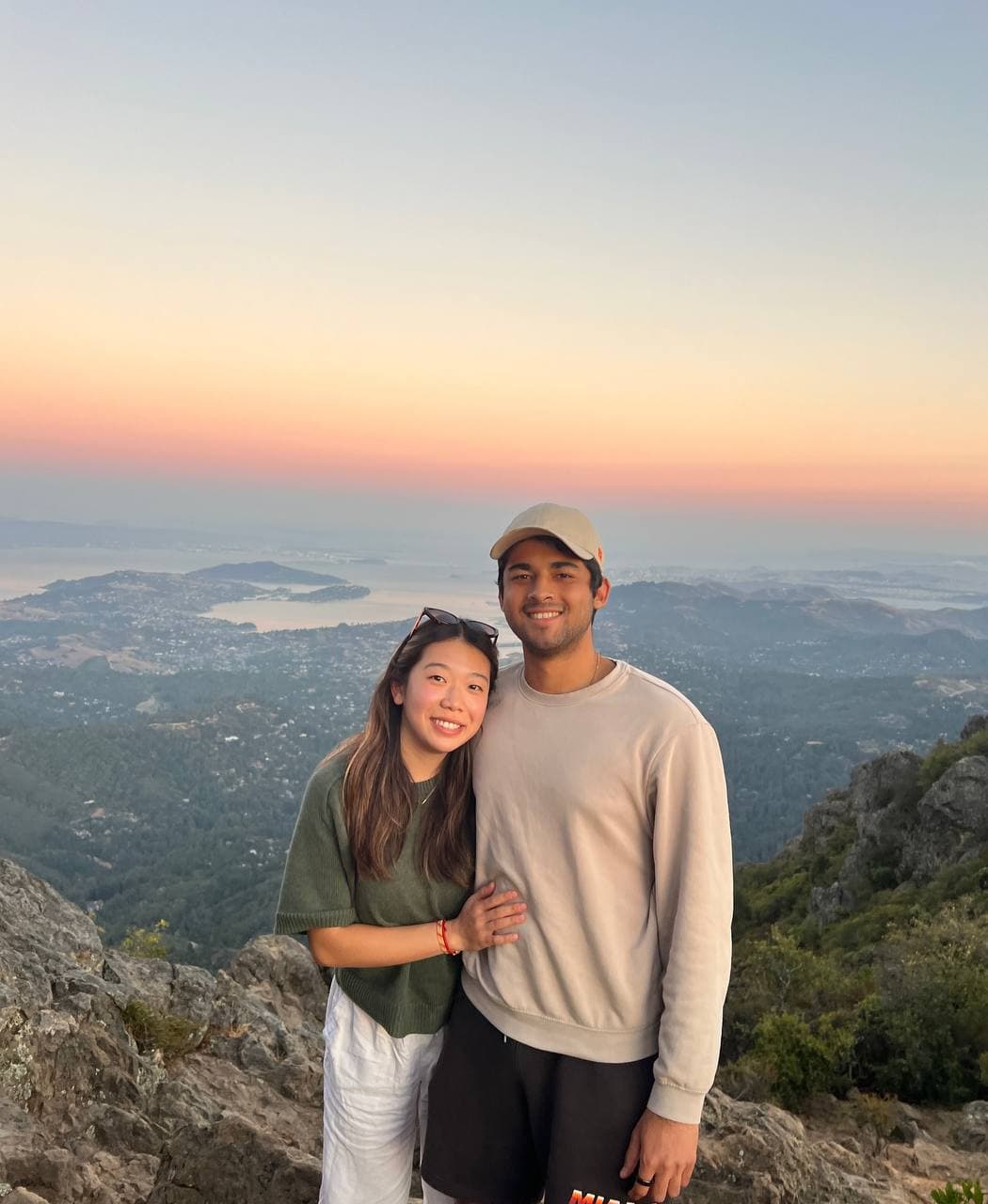 Amy in a green top and white pants with arms around Roshan on a mountaintop, sun setting over the bay