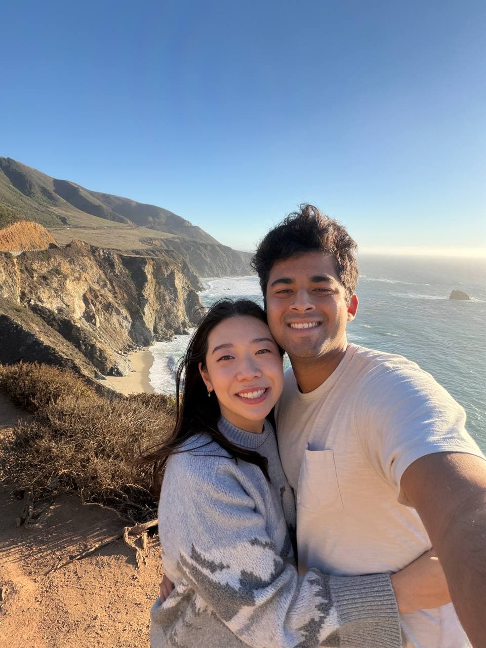 Amy and Roshan smiling on a Big Sur cliffside trail, rugged coastline behind them