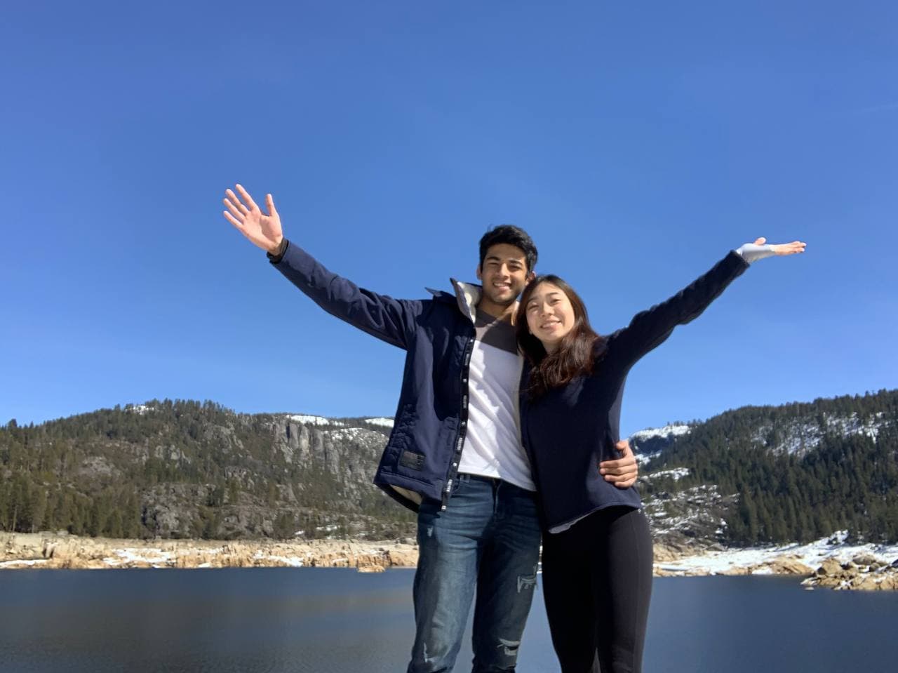 Amy and Roshan with arms raised at a mountain lake