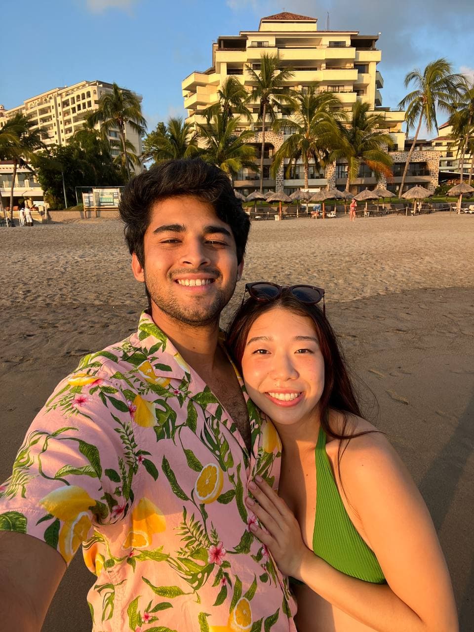 Roshan in a pink lemon-print Hawaiian shirt with Amy, smiling on a beach at sunset